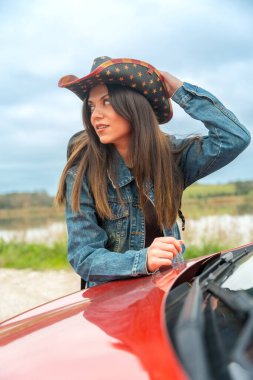 Yolun ortasında kırmızı bir arabaya yaslanan dikkati dağılmış bir gezginin dikey portresi.