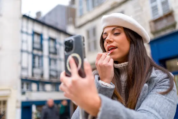 Chic woman with beret making up in the street using the mobile as a mirror