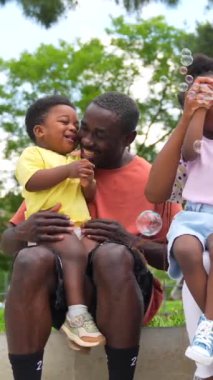Parktaki ağaçların yanında sabun köpüğü üfleyerek eğlenen Afrikalı siyahi etnik aile.