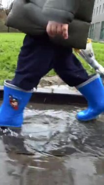 Mavi lastik çizmeli bir çocuğun ayakları yağmurda su birikintisinin üzerinden atlıyor.