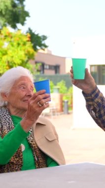 Huzurevinin bahçesinde neşeli iki son sınıf öğrencisi, güneşli bir yaz gününde canlı bardaklarla kadeh kaldırıyorlar.