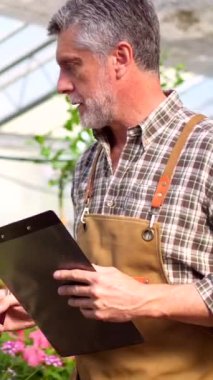 Bahçıvan çiçek serasının içindeki bir çocuk odasında çalışıyor, bir klasörle envanter çıkarıyor.