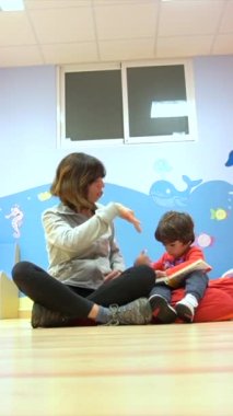 Çocuklu bir kadın öğretmen oturmuş kitap okuyor ve bir anaokulunda anlatılanlarla gülüyor.