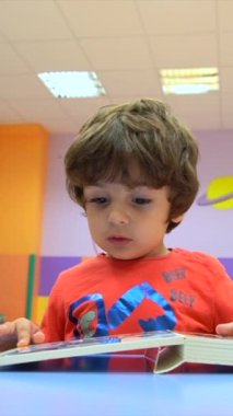 Masada oturmuş masal kitabı okuyan bir çocuğun portresi. Bir anaokulunun içinde kameraya bakıyor.