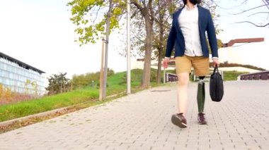 Şehir parkı boyunca dizüstü bilgisayar çantası taşıyan protez bacaklı bir iş adamının ön görüntüsü.