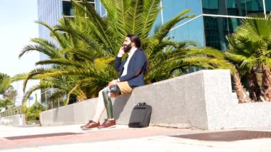 Protez bacaklı işadamı gülümseyen cep telefonuyla konuşuyor. Finans binasının önünde oturuyor.