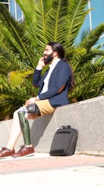 Protez bacaklı işadamı gülümseyen cep telefonuyla konuşuyor. Finans binasının önünde oturuyor.