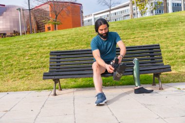 Fiziksel engelli erkek koşucu, parkta bir bankta oturarak protez bacağını düzeltiyor.