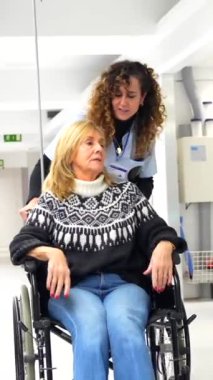 Hemşire hastane koridorunda tekerlekli sandalyede yetişkin bir kadını taşıyor.
