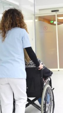 Hastane koridorunda tekerlekli sandalyeyi iten bir hemşirenin arka görüntüsü.