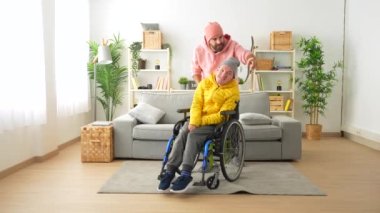 Bir bakıcı portresi ve tekerlekli sandalyede sakat bir adam evde kameraya bakıp gülümsüyor.