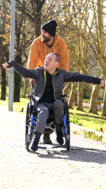 Tekerlekli sandalyede oturan engelli bir adam ve doğanın tadını çıkaran bir arkadaşı parkı işaret edip etrafa bakınıyorlar.