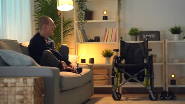 車椅子の隣のソファーに座って絶望を眺めながら自宅で一人で悲しい障害者 — ストック動画