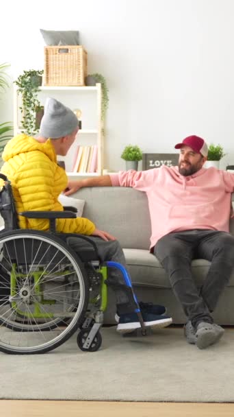 自宅でソファーに座っている間 車椅子で障害のある友人をサポートする男 — ストック動画