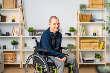 Modern bir dairenin oturma odasında gülümseyen tekerlekli sandalyedeki mutlu engelli adam.
