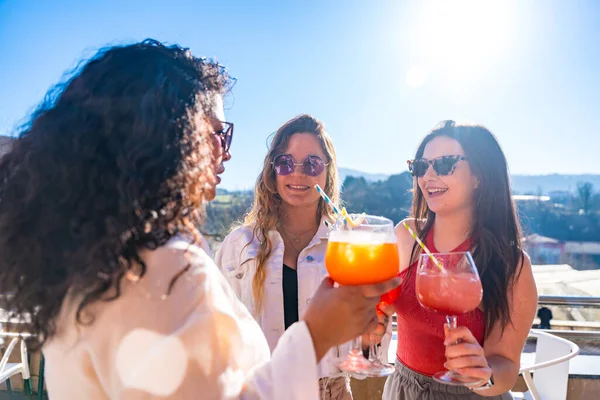 Three cool female friends enjoying summer toasting with cocktails on a rooftop in a sunny day