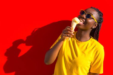 Gülümseyen ve şakacı Afrikalı kız kırmızı duvarda dondurma yiyor.