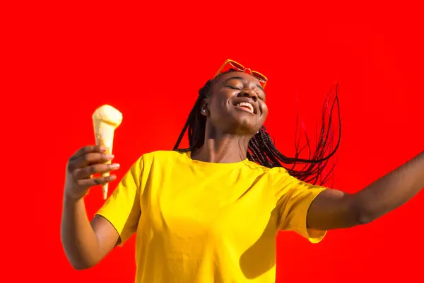 Happy african woman dancing while eating ice cream against red background