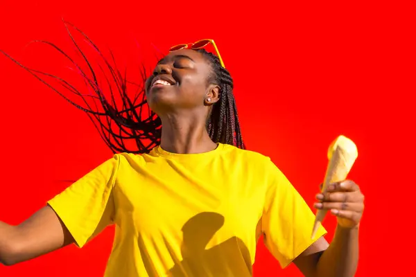 Cool happy african woman dancing and eating ice cream under the sun and against a red wall