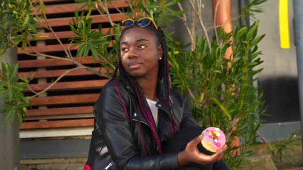 Αφρικανική Χαριτωμένο Γυναίκα Τρώει Ένα Πολύχρωμο Ντόνατ Κάθεται Εξωτερικούς Χώρους — Αρχείο Βίντεο