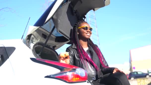 在一个阳光灿烂的日子里 坐在汽车后备箱里享用甜甜圈的非洲女人 — 图库视频影像