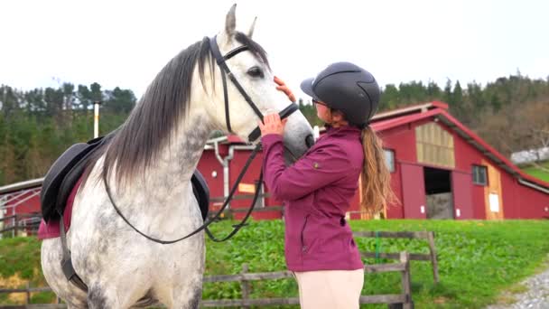 馬の頭を馬の中心に世話するヘルメットを持つ女性の若いジョッキー — ストック動画