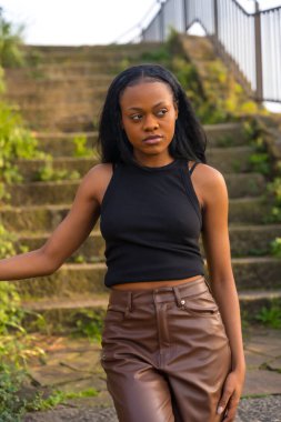 Afrikalı bir kadın şehir parkında merdivenli şık kıyafetlerle poz verirken başka tarafa bakıyor.