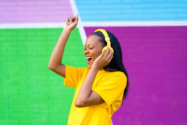 Happy african woman dancing and listening to music using headphones next to a urban multicolored wall