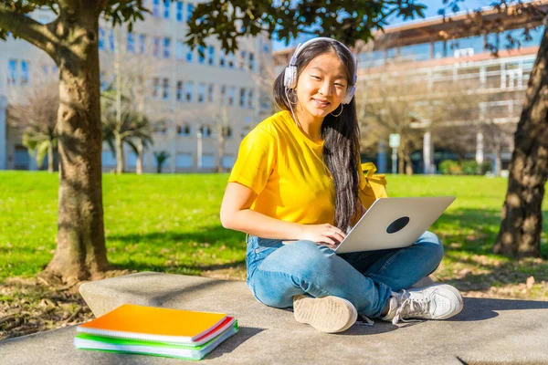Smiling chinese student looking at camera while studying with laptop and headphones outdoors