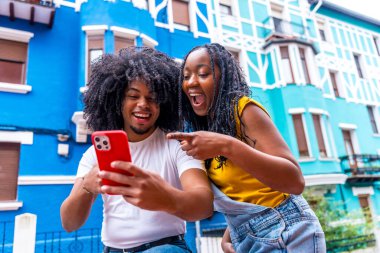 Afrikalı arkadaşlar, şehirde mavi renkli evlerin yanında duran, şaşırmış bir ifadeye sahip bir cep telefonunu işaret ediyorlar.