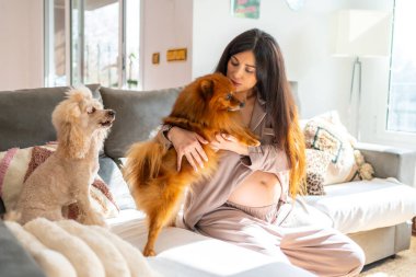 Hamile bir kadın evdeki kanepede oturmuş küçük köpeklerini öpüyor ve okşuyor.