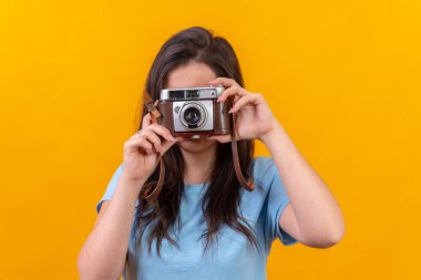 Eski moda bir fotoğraf makinesi kullanan sıradan bir genç kadının sarı arka planlı stüdyo portresi.