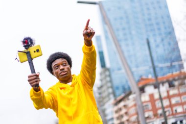 African young vlogger pointing ahead during an online video with a mobile attached on mic and tripod in the city clipart