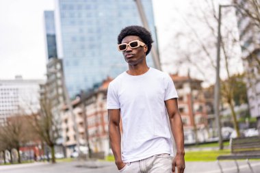 Beyaz elbiseli, güneş gözlüklü, havalı ve genç Afrikalı bir adam şehirde yürüyor.