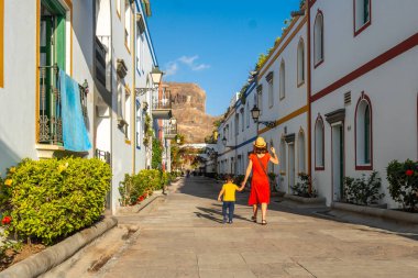 Kırmızı elbiseli bir anne ve oğlu Gran Canaria 'daki Mogan kasabasının limanında yürüyor. Aile tatili