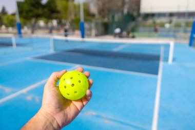 Arka planda mavi kortlu plastik sarı bir picleball topu tutan tanınmamış bir kişi.