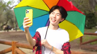 Parkta elinde gökkuşağı renkli bir şemsiye tutarken cep telefonuyla selfie çeken Çinli eşcinsel bir adam.
