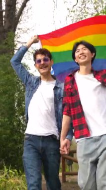 Çok ırklı eşcinsel bir çiftin parkta lgbt bayrağı çekerken düşük açılı portresi.