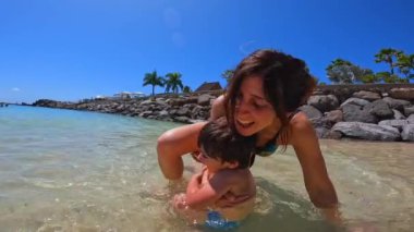 Bir kadın ve bir çocuk okyanusta oynuyorlar. Kadın, çocuğun suyun içinde ayakta durmasına yardım ediyor. Manzara neşeli ve eğlenceli.