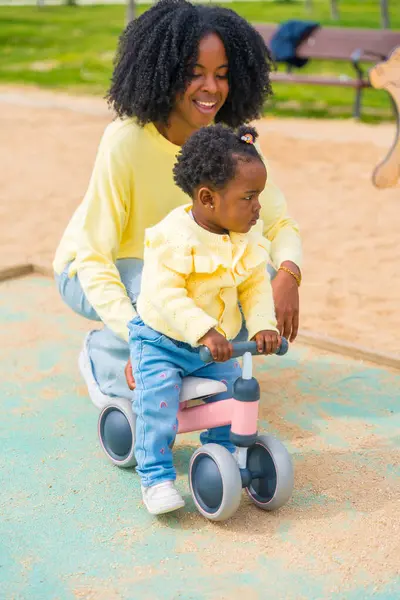 Little african girl learning to ride a bike with her mother in a public playground