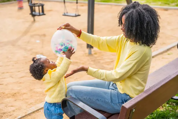 非洲母亲和女儿坐在公园的长椅上玩气球 — 图库照片