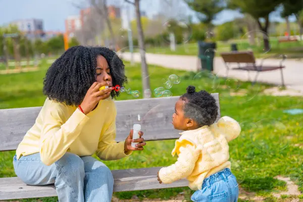 非洲母亲和女儿坐在公园的长椅上玩肥皂泡 — 图库照片