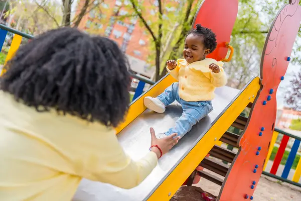 公共の遊び場でスライドで彼女の母親と遊ぶ幸せなアフリカの子供 — ストック写真