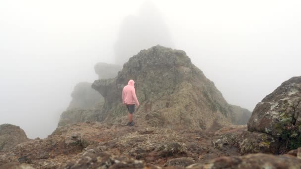 一个穿着粉色帽衫的男人正在爬上一座岩石山 天空阴沉沉的 天气多云 免版税图库视频