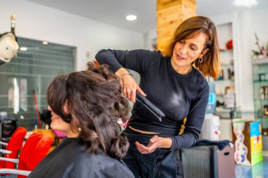 Kuaför ve müşteri bir güzellik salonunda saçlarını kıvırırken konuşuyor.