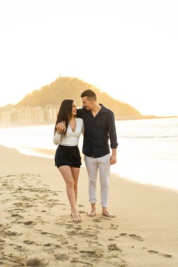 Gün batımında sahilde çıplak ayakla yürüyen romantik bir çiftin dikey fotoğrafı.