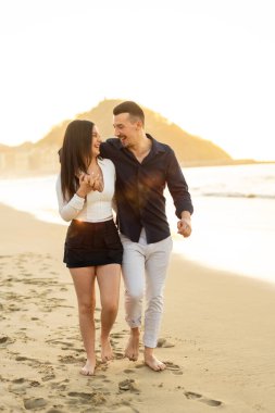 Gün batımında sahilde çıplak ayakla yürüyen romantik bir çiftin dikey fotoğrafı.