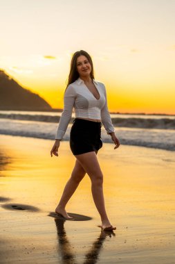 Gün batımında plajda çıplak ayakla yürüyen zarif, şehvetli bir kadının dikey portresi.