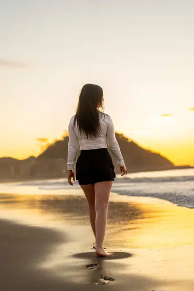 日落时赤脚在海滩上行走的性感优雅女子的垂直后视镜 图库图片