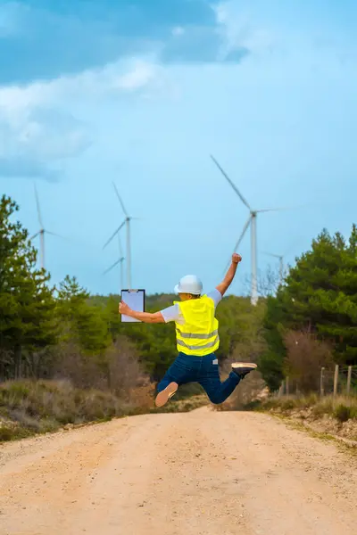 Vertical rear view of a male caucasian adult engineer celebrating jumping on air next to wind turbines in a park